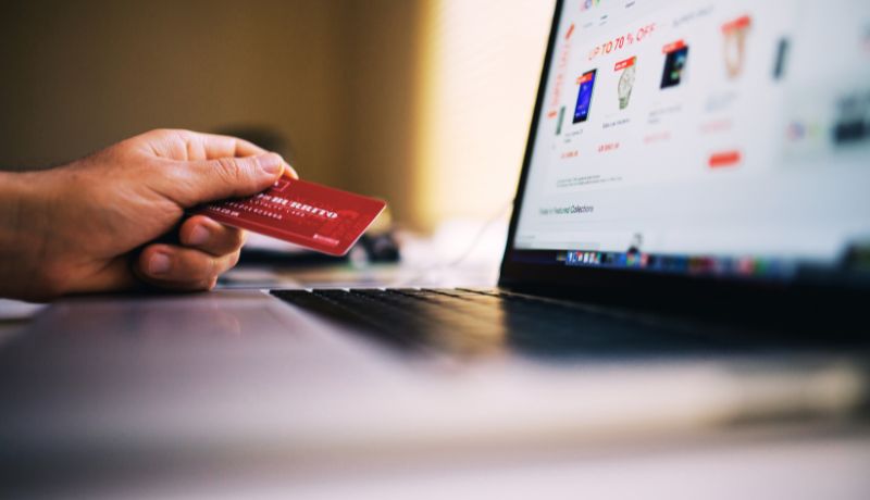 A close-up of a hand holding a red credit card in front of a laptop screen displaying an online shopping website with various products and discounts.