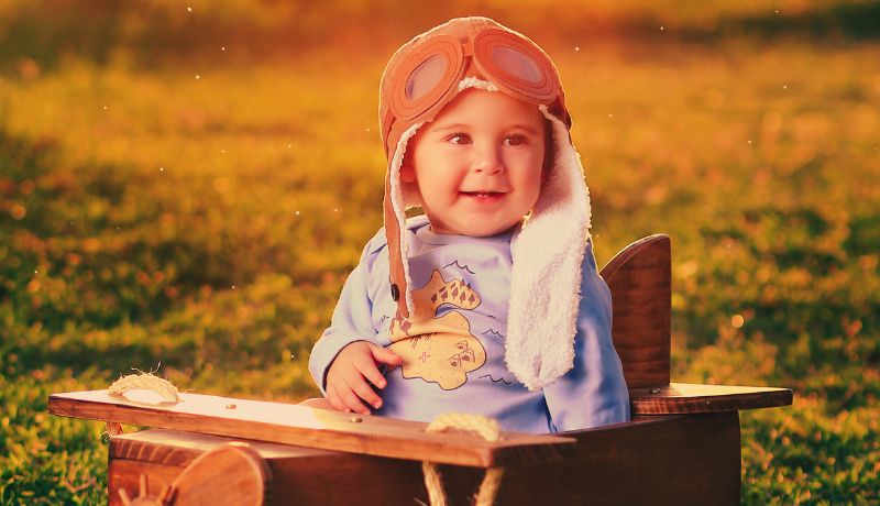 A young child sitting in a wooden toy airplane in an aviator hat with goggles on a grassy field with a warm, golden light,