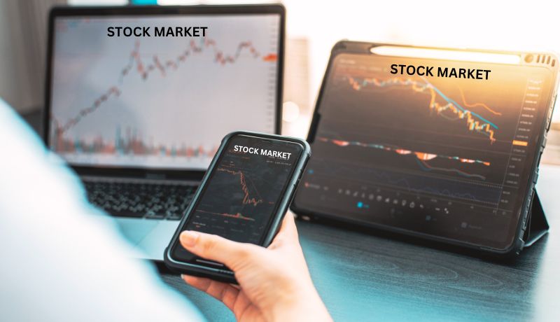 The image shows three electronic devices displaying stock market charts. There is a laptop in the background, a tablet to the right, and a smartphone being held in a hand in the foreground.