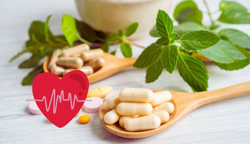Two wooden spoons filled with capsules and surrounded by green leaves, and also a red heart icon with a white heartbeat line superimposed on the image